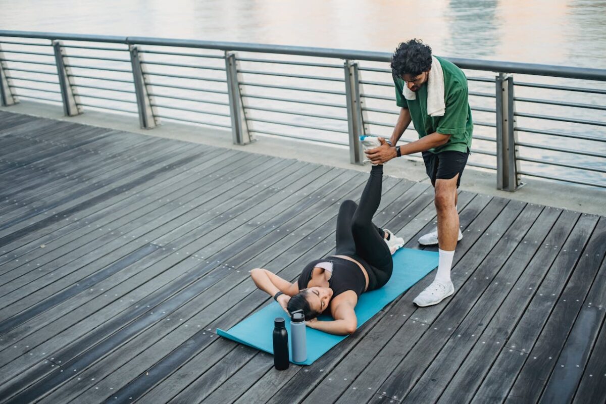 Man assisting woman in stretching exercises on boardwalk next to river, emphasizing fitness and teamwork.