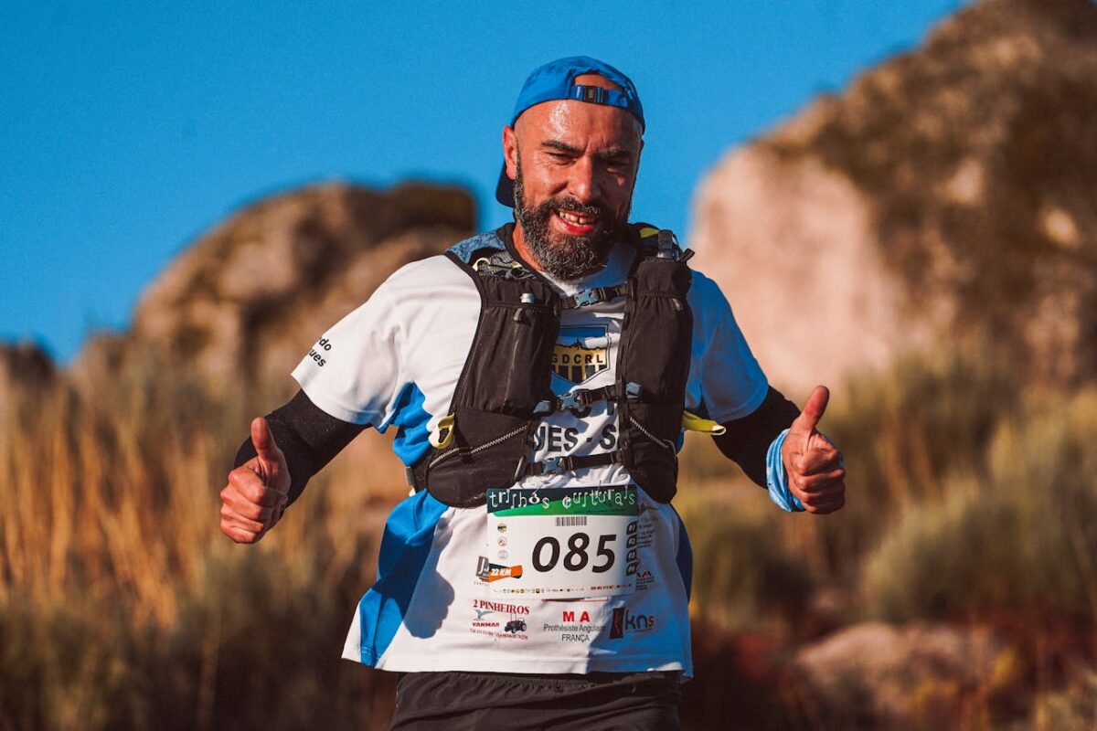 Smiling man wearing sports gear gives thumbs up during an outdoor trail race, exuding joy and energy.